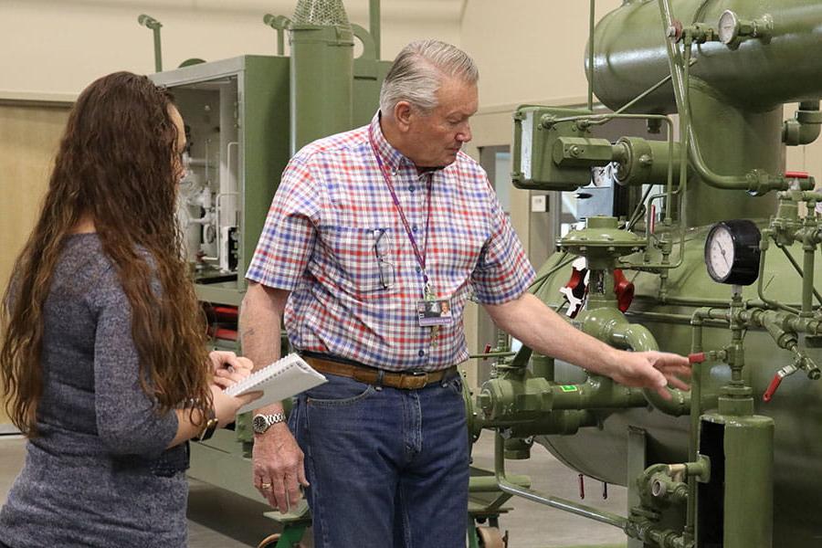 An individual touches valves on machinery while another individual looks on and takes notes in a notebook